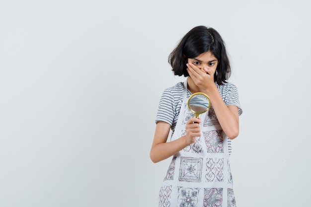 Menina olhando para a lente de aumento de camiseta, avental e parecendo surpresa,