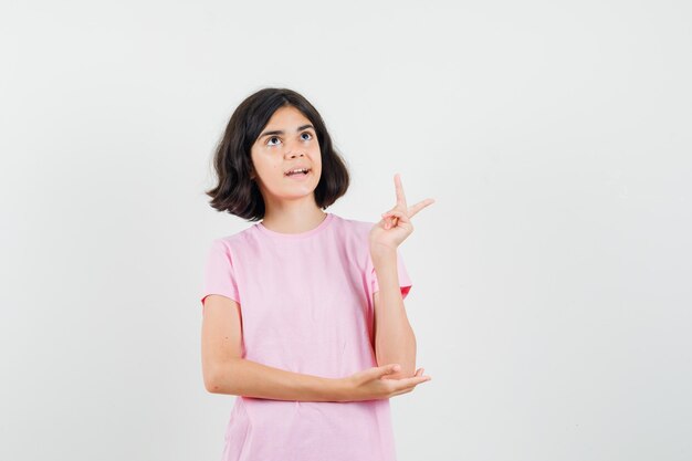 Menina olhando para cima, mostrando o sinal V em uma camiseta rosa, vista frontal.