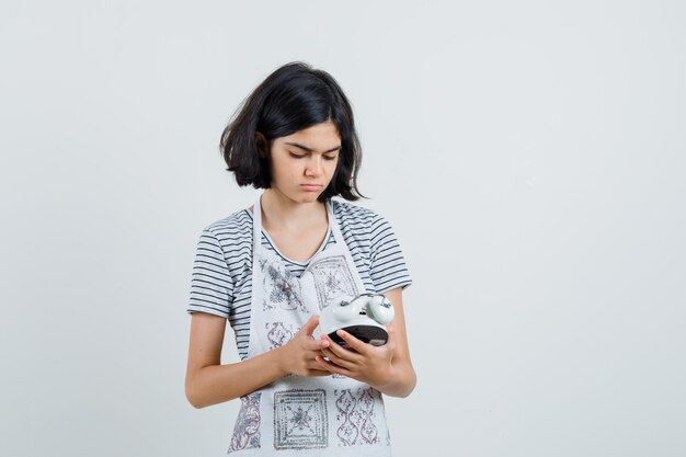 Menina olhando para o despertador em t-shirt, avental e olhando pensativa.