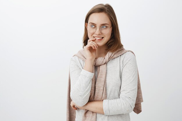 Menina olhando para um cara curioso tentando desviar a atenção, mordendo o dedo e sorrindo intrigada olhando para a esquerda com interesse e desejo, tendo uma ideia misteriosa e deliciosa em pé sobre uma parede cinza