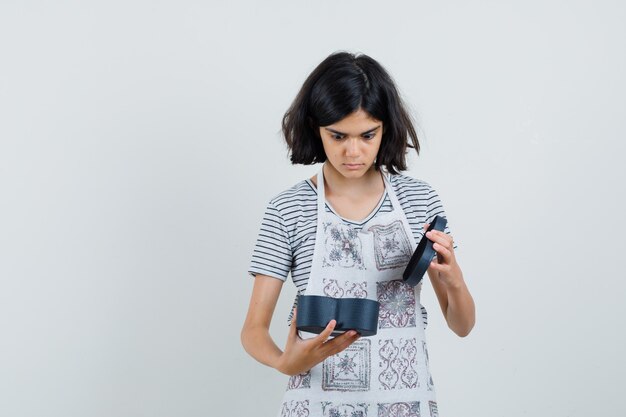 Menina olhando para uma caixa de presente em t-shirt, avental e parecendo surpresa,