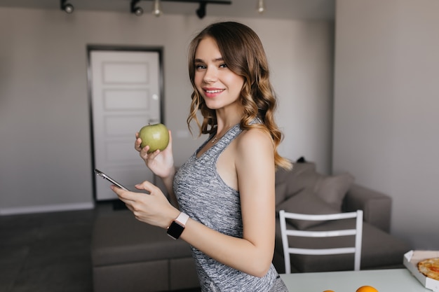 Foto grátis menina pálida magro em smartwatch posando em casa. senhora encantadora de cabelos escuros com maçã se divertindo pela manhã.