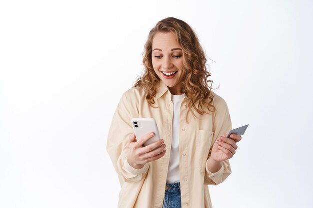 Menina parece feliz na tela do smartphone e sorri, segura um cartão de crédito de plástico, faz um pedido e fica em pé sobre uma parede branca