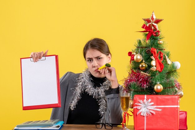 Menina pensativa de vista frontal sentada à mesa segurando um documento com a árvore de natal da coroa e coquetel de presentes