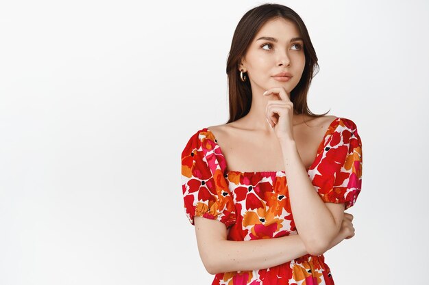 Menina pensativa elegante em vestido floral feminino olhando para o canto superior direito e pensando em fazer a escolha em pé sobre fundo branco