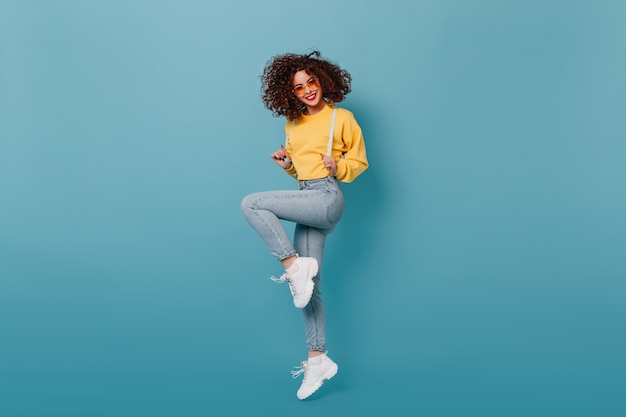 Menina positiva com batom vermelho vestida com moletom amarelo e calça jeans skinny pulando no espaço azul. instantâneo de senhora encaracolada em óculos laranja.