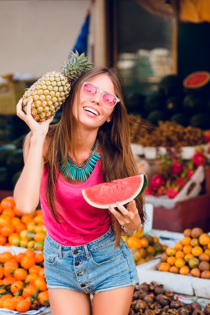 Menina positiva com um grande sorriso segurando ananás e uma fatia de melancia no mercado