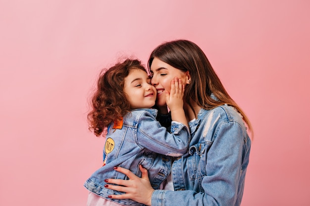 Menina pré-adolescente relaxada, abraçando-se com a mãe. Winsome jovem mãe beijando filha no fundo rosa.