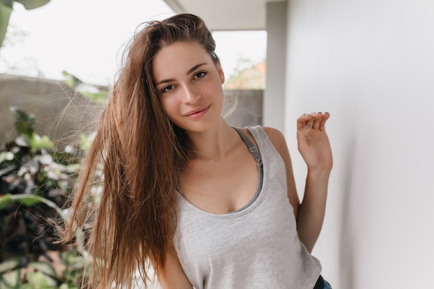 Menina preguiçosa de cabelos compridos, posando com expressão facial interessada. Foto ao ar livre de uma deslumbrante mulher morena em pé perto de uma parede branca de manhã.