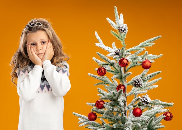 Menina preocupada em pé perto da árvore de natal, usando uma tiara com uma guirlanda no pescoço, colocando as mãos nas bochechas isoladas em um fundo laranja