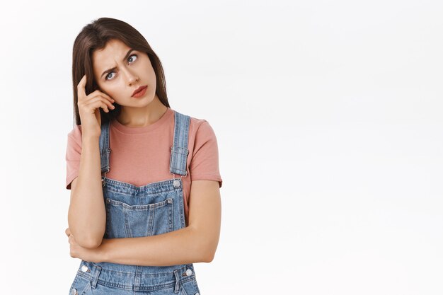 Menina preocupada em resolver quebra-cabeças, parada confusa e pensativa, inclinar a cabeça, franzir a testa e coçar a cabeça pensando, parada pensativa, tentando tomar uma decisão difícil ou encontrar uma solução, fundo branco