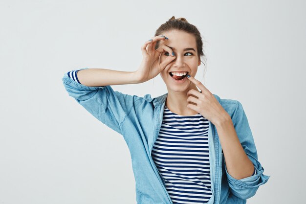 Menina pronta para descobrir novos horizontes. Alegre mulher bonita europeia segurando o gesto bem sobre o olho e olhando de lado através dele, tocando o rosto com o dedo indicador e estar de bom humor