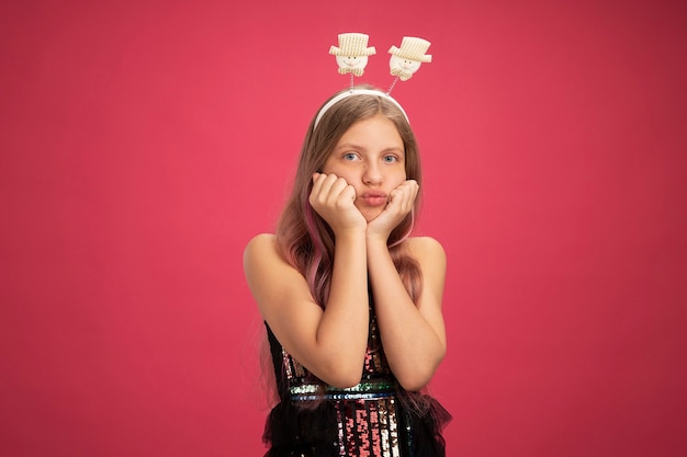 Menina ressentida com vestido de festa brilhante e tiara engraçada olhando para a câmera soprando bochechas conceito de feriado de celebração de ano novo em pé sobre fundo rosa