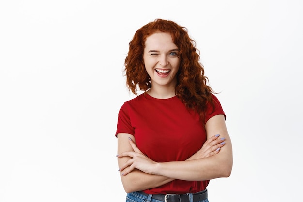 Menina ruiva animada com cabelo encaracolado vestindo camiseta piscando e sorrindo feliz em pé com os braços cruzados sobre o peito permanecendo positivo contra o fundo branco