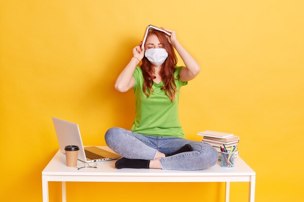 Menina ruiva com máscara médica se senta com as pernas cruzadas na mesa branca com o livro acima da cabeça, mantém os olhos fechados, vestindo jeans e camiseta verde, rodeada de lap top, café, canetas.