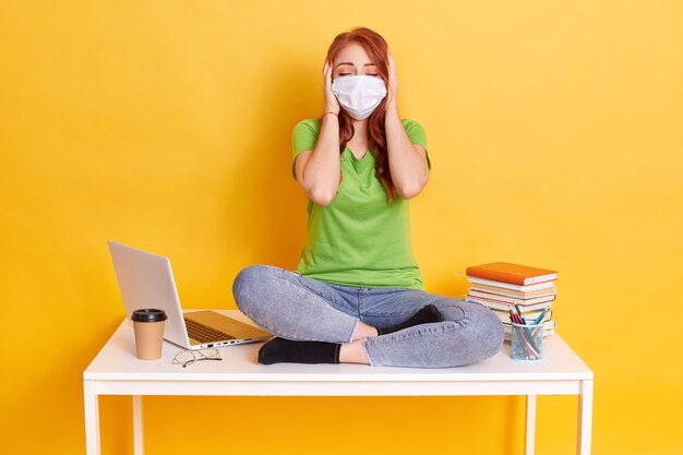 Menina ruiva com máscara médica sentada com as pernas cruzadas na mesa branca, cobrindo as orelhas com as palmas das mãos e mantendo os olhos fechados, vestindo jeans e camiseta, rodeada de lap top, café, canetas