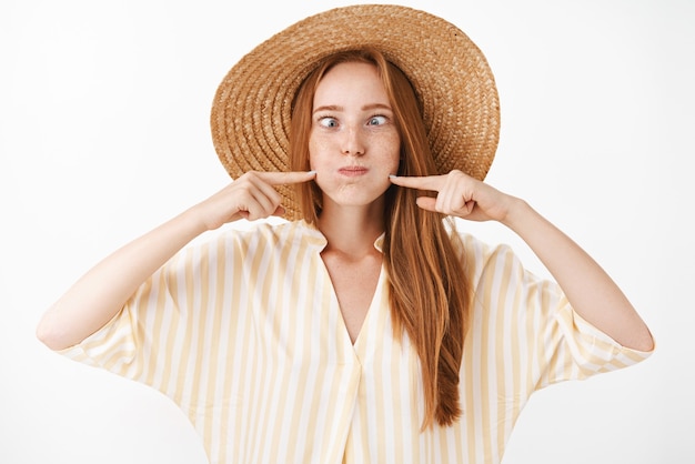 Menina ruiva divertida e alegre, bonita, despreocupada, com sardas fazendo caretas engraçadas, segurando a respiração e fazendo beicinho cutucando as bochechas com os dedos para soprar, olhando para o nariz