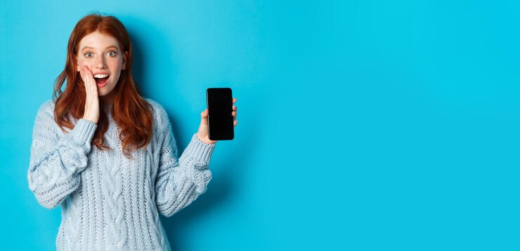 Foto grátis menina ruiva espantada olhando para a câmera mostrando a tela do smartphone demonstrando a aplicação móvel sta