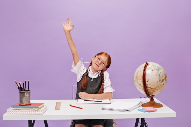 Foto grátis menina ruiva inteligente com tranças e sardas na blusa branca e vestido de escola cinza, levantando a mão no pano de fundo lilás