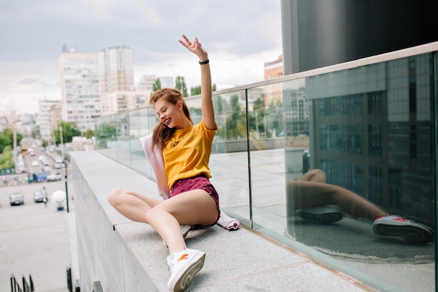 Menina ruiva sorridente com camisa amarela deitada no parapeito de pedra e olhando para baixo