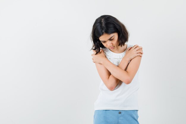 Menina se abraçando em uma blusa branca, shorts e parecendo triste, vista frontal.