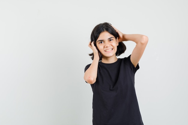 Menina segurando a cabeça com as mãos em uma camiseta preta e olhando feliz, vista frontal.