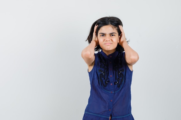 Menina segurando a cabeça com as mãos na blusa azul e parecendo irritada, vista frontal.