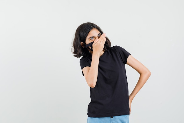 Menina segurando a mão na boca em camiseta preta, shorts, máscara e parece feliz. vista frontal.