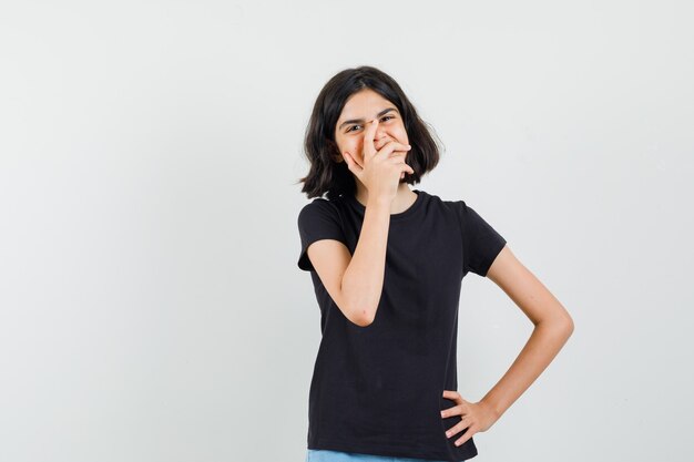 Menina segurando a mão na boca em t-shirt preta e parecendo feliz. vista frontal.