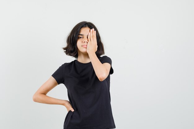Menina segurando a mão no olho em t-shirt preta, vista frontal.
