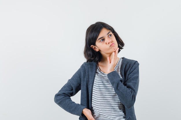 Menina segurando a mão no queixo com camiseta, jaqueta e parecendo hesitante