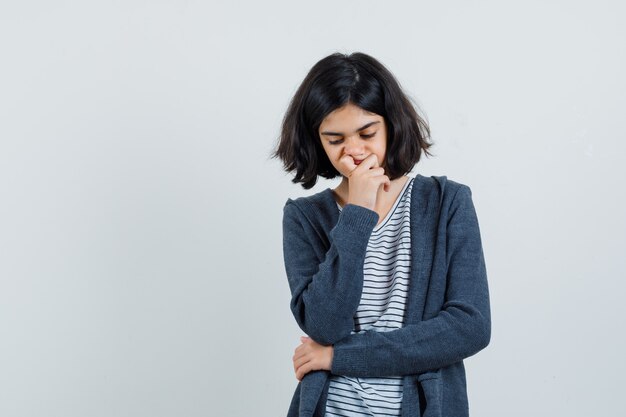 Menina segurando a mão no queixo com camiseta, jaqueta e parecendo pensativa,