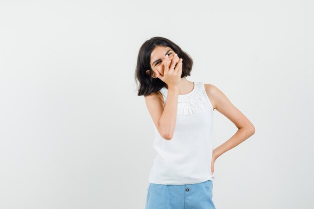 Menina segurando a mão no rosto de blusa branca, shorts e olhando feliz, vista frontal.