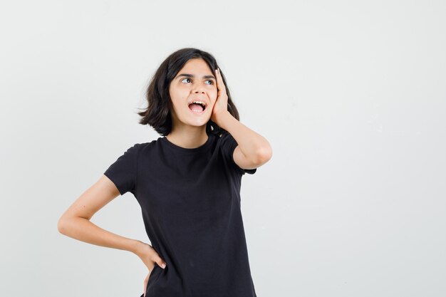 Menina segurando a mão no rosto em t-shirt preta e olhando esquecido, vista frontal.