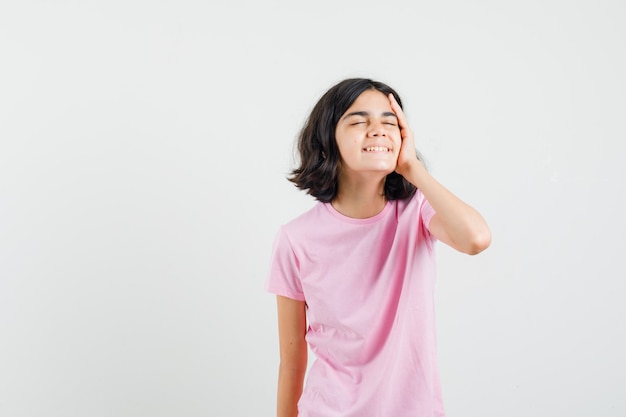 Menina segurando a mão no rosto em t-shirt rosa e olhando alegre, vista frontal.