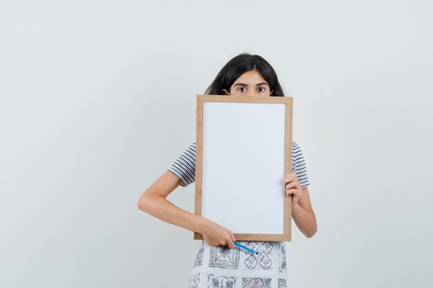 Menina segurando moldura branca, lápis na t-shirt, avental e parecendo perplexa.