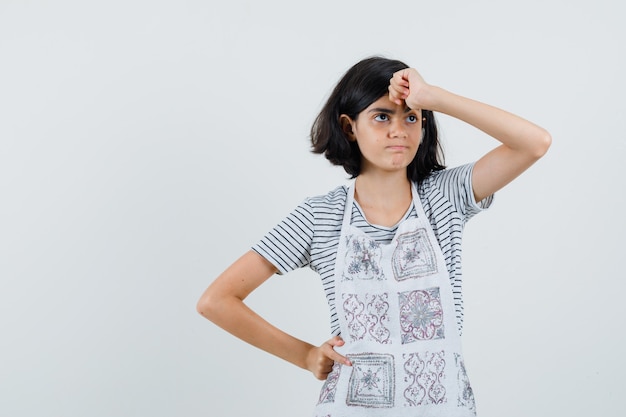 Menina segurando o punho na testa com camiseta, avental e parecendo arrependida,