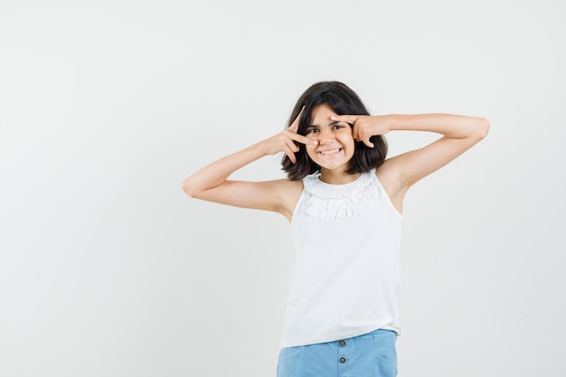 Menina segurando os dedos no rosto em blusa branca, shorts e bonito. vista frontal.