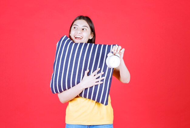 Menina segurando um travesseiro azul com listras brancas e mostrando um despertador.