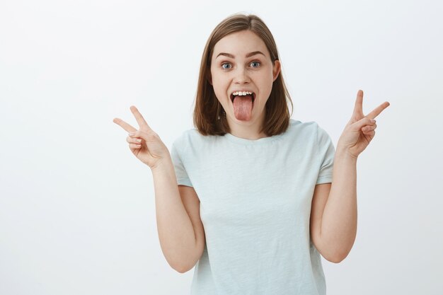 Menina sem medo de se expressar. Mulher alegre e brincalhona feliz confiante em uma camiseta casual mostrando a língua, os sinais da paz com as duas mãos brincando e se divertindo desfrutando da juventude sobre a parede cinza