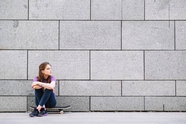 Menina sentada em um skate contra uma parede de tijolos