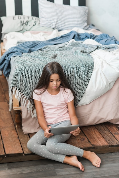 Foto grátis menina sentada perto da cama olhando para tablet digital