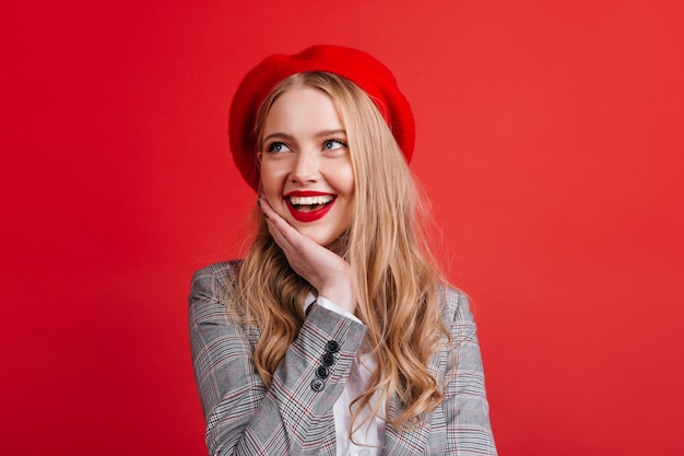 Menina sonhadora caucasiana com cabelo loiro, olhando para cima com um sorriso. mulher francesa positiva na boina isolada na parede vermelha.
