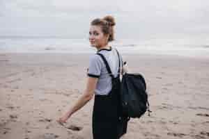 Foto grátis menina sonhadora com mochila preta olhando por cima do ombro enquanto caminhava ao longo da costa. foto ao ar livre da alegre modelo feminina branca, passando um tempo na praia.