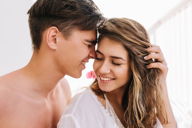 Menina sonhadora com sorriso animado, segurando o cabelo dela e ouvindo a confissão de amor de um jovem bonito com cabelo curto escuro. Retrato de menino e a namorada passando um tempo juntos pela manhã