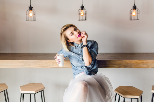 Menina sonhadora e elegante, com cabelo loiro e lábios cor de rosa, sentada em um café com cadeiras de madeira e mesa. ela segura uma xícara de café