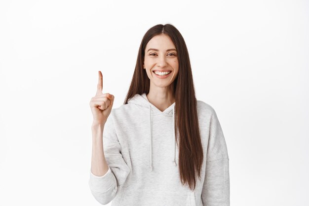 Menina sorridente alegre com sorriso perfeito e rosto limpo, apontando o dedo para cima no logotipo superior, mostrando o banner de anúncio acima de pé em fundo branco com capuz casual