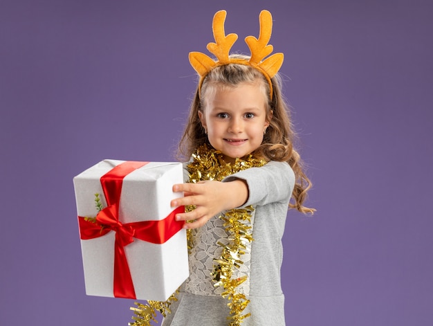 Menina sorridente com aro de cabelo de natal e guirlanda no pescoço segurando uma caixa de presente isolada na parede azul