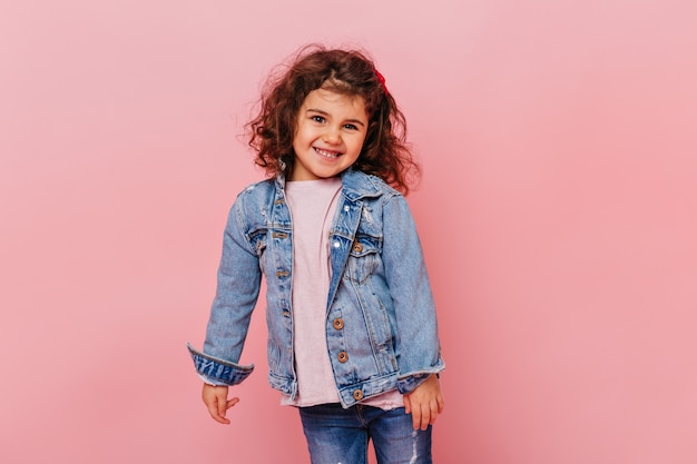 Menina sorridente com cabelo ondulado em pé sobre fundo rosa. Foto de estúdio de adorável criança pré-adolescente vestindo jaqueta jeans.