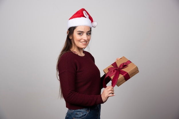 Menina sorridente com chapéu de papai noel, segurando a caixa de presente em fundo cinza.